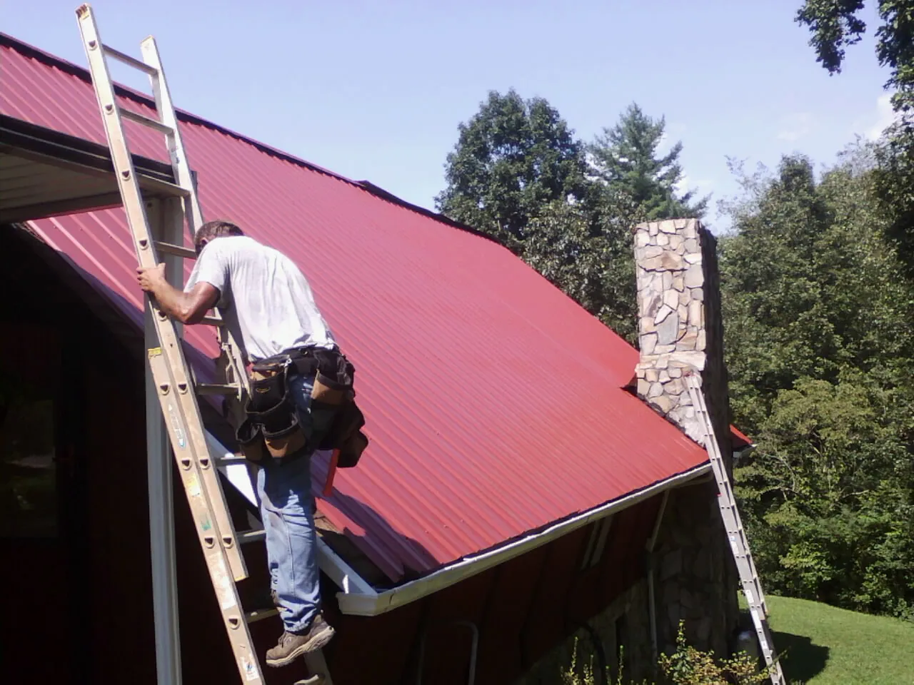 Roofer on a ladder beside a red metal roof and stone chimney — Soffit & Fascia Repair by Roofing Brothers in Chinook